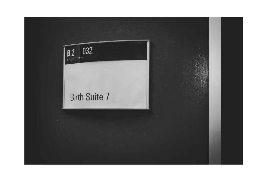 Black-and-white photo of a hospital door sign that reads “Birth Suite 7”.