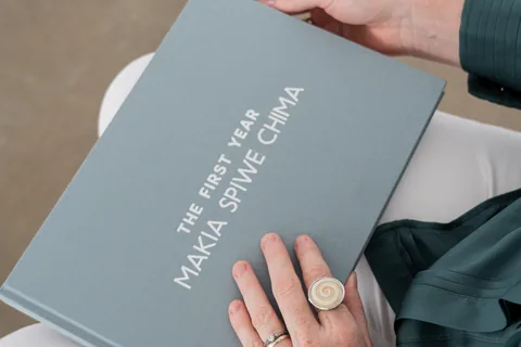 The optional printed hardcover: a finished First Year journal held on a lap, in grey linen with the baby's name on the cover