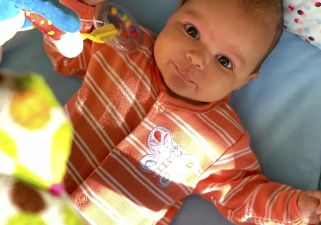 A photo of the baby at three months, smiling up from a play gym in an orange striped romper