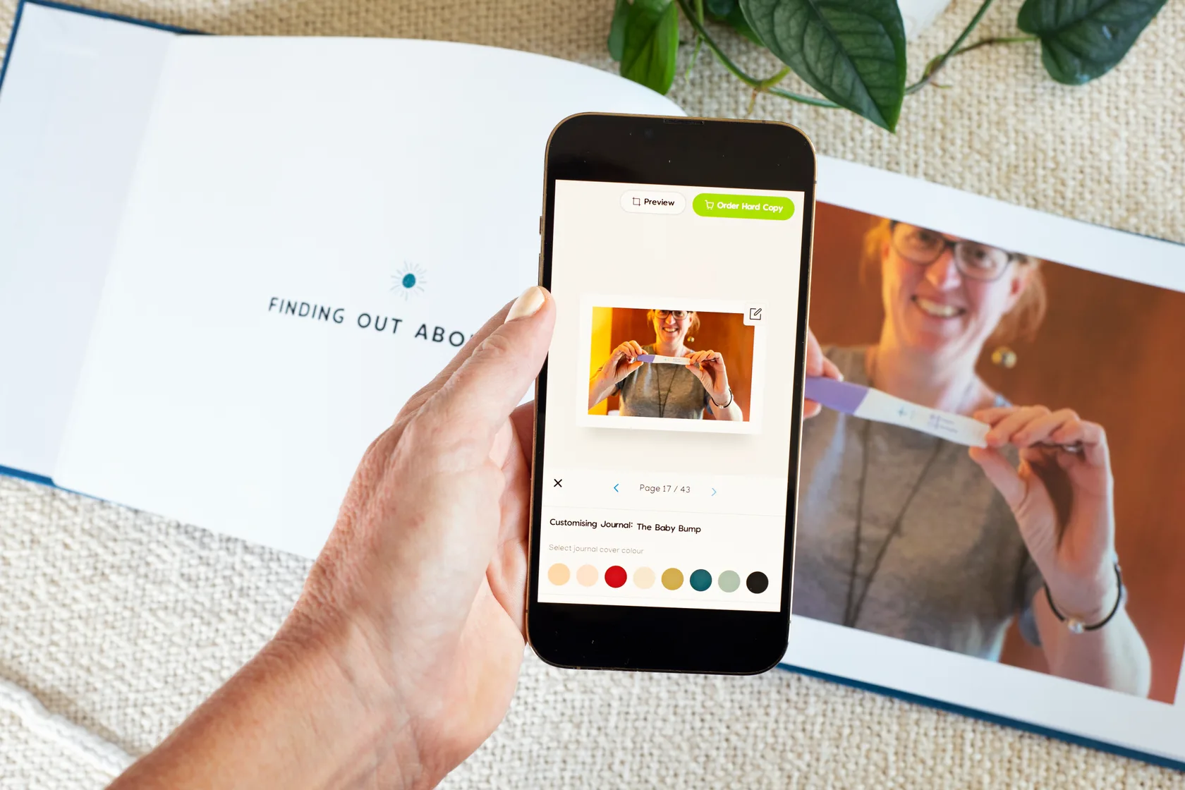 A hand holds a phone showing the Makia app customising a Baby Bump journal, beside the same memories printed in an open linen keepsake on the couch