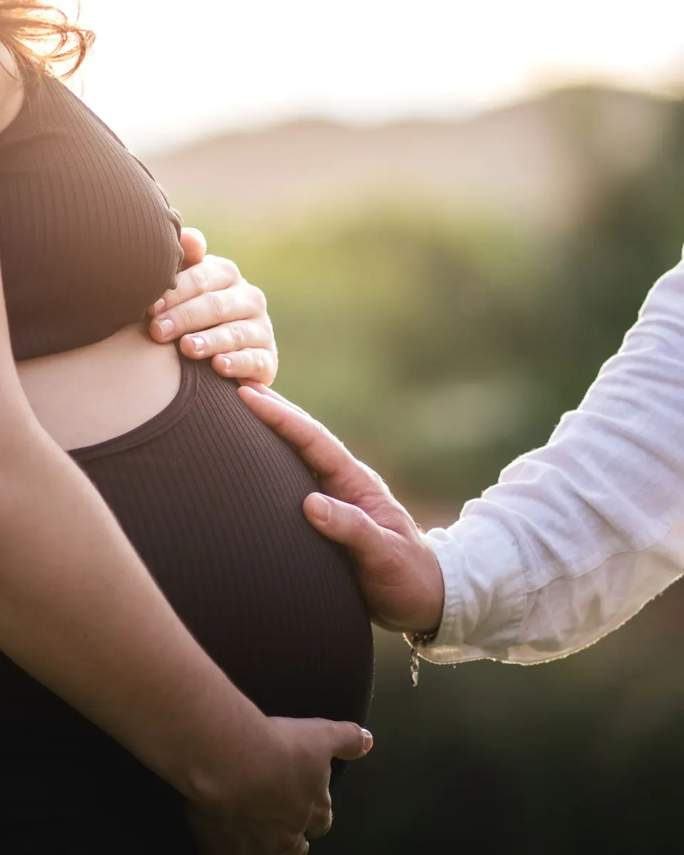 A partner's hand resting on a pregnant belly in soft evening sunlight, beside the parent's own hands.