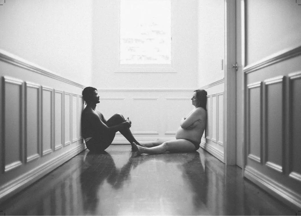 Black-and-white full-page portrait of expecting parents sitting face to face in a hallway.