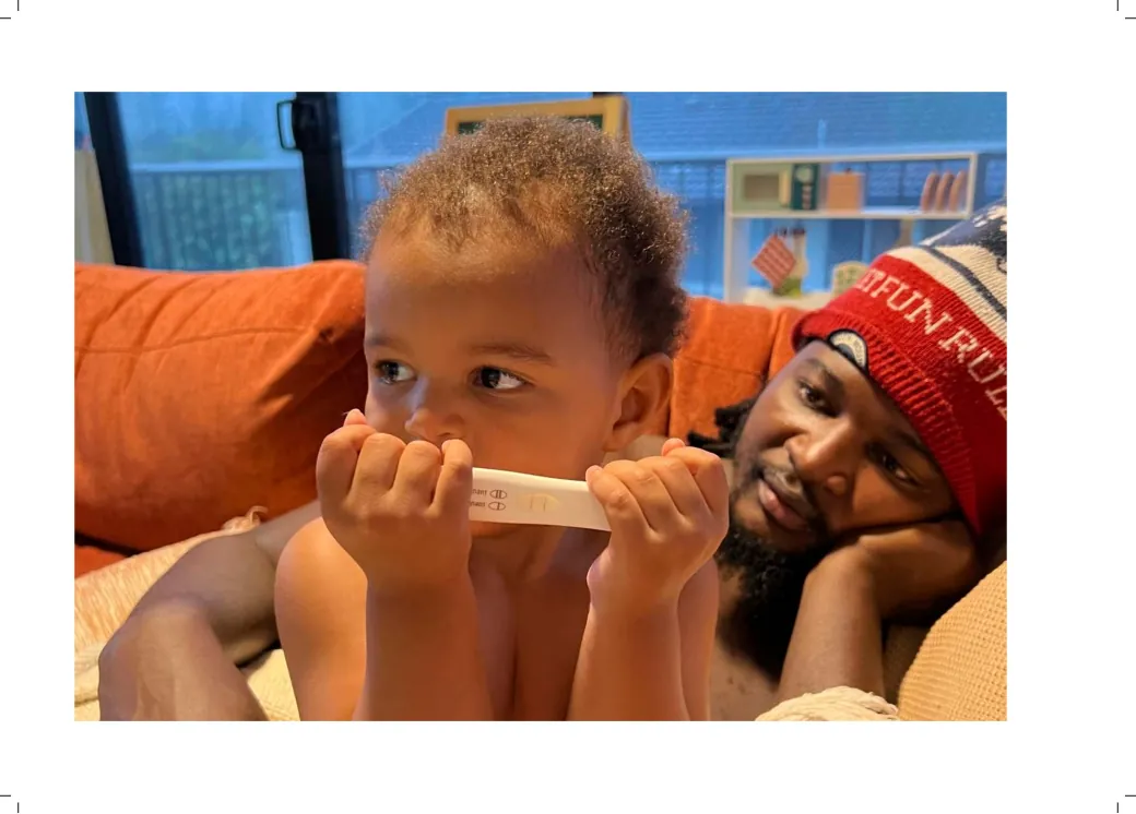 Full-page photo of a toddler holding a positive pregnancy test, their dad resting beside them.
