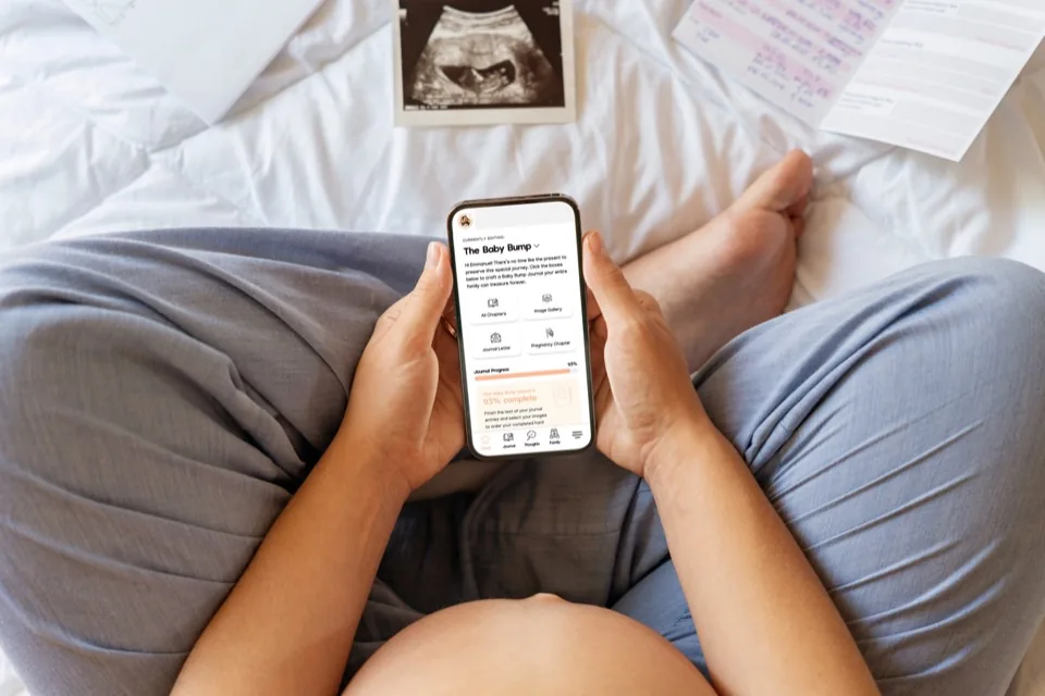 A pregnant parent using the journal on a phone, with an ultrasound print on the bed.